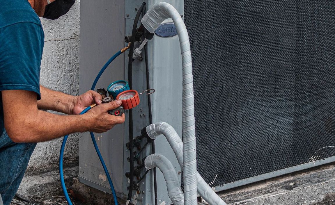 Technician repairing an air conditioner unit outdoors, wearing a facemask and using a manifold gauge.